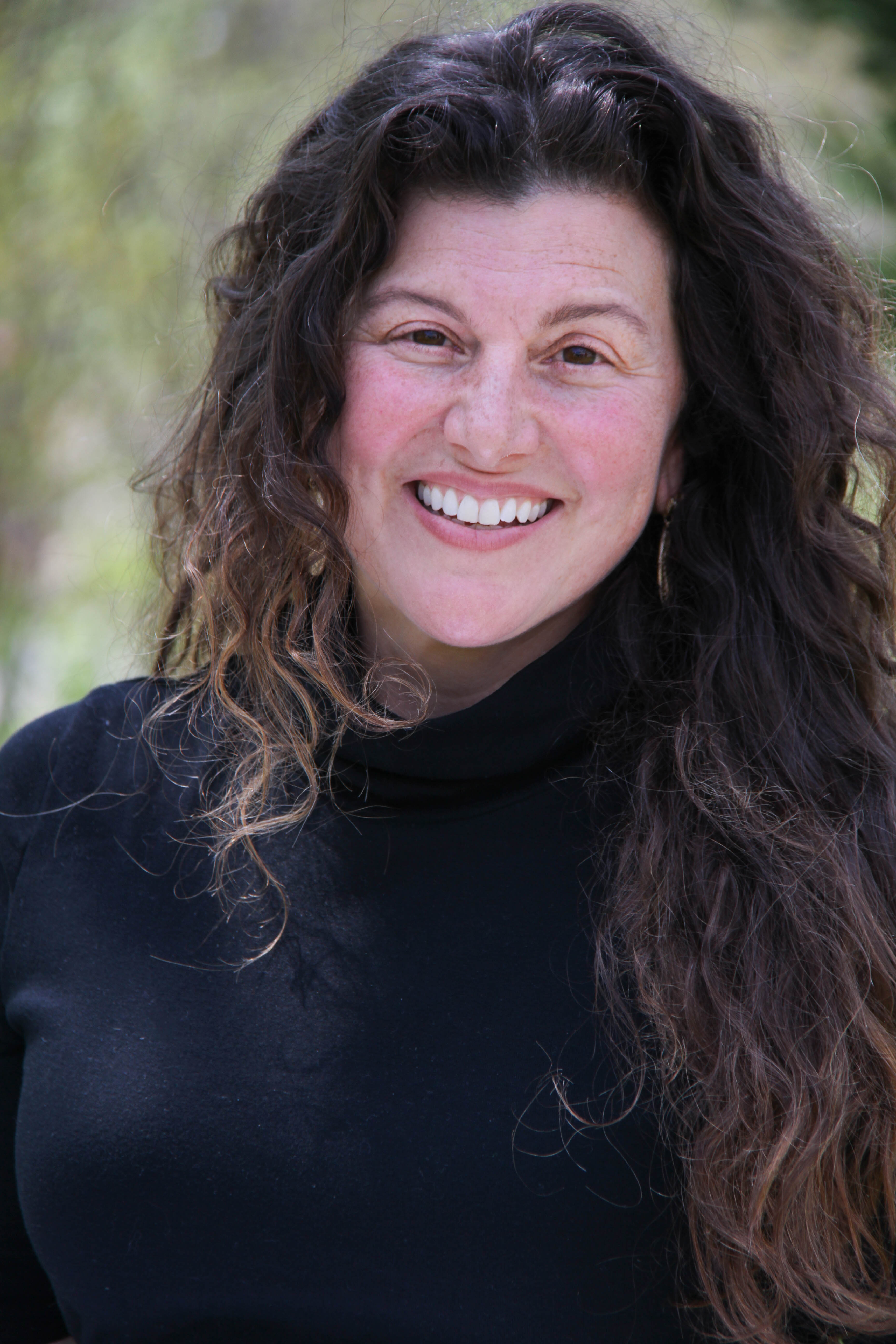 Lisa Condino smiles at the camera. Her long, brown, curly hair drapes and swirls around her face and over her shoulders. She wears a black sweater.