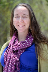 Alexandra Turner smiles widely at the camera, with long, brown hair draped over her shoulders. She wears a cobalt blue dress, green sweater, and purple scarf with white polka dots.