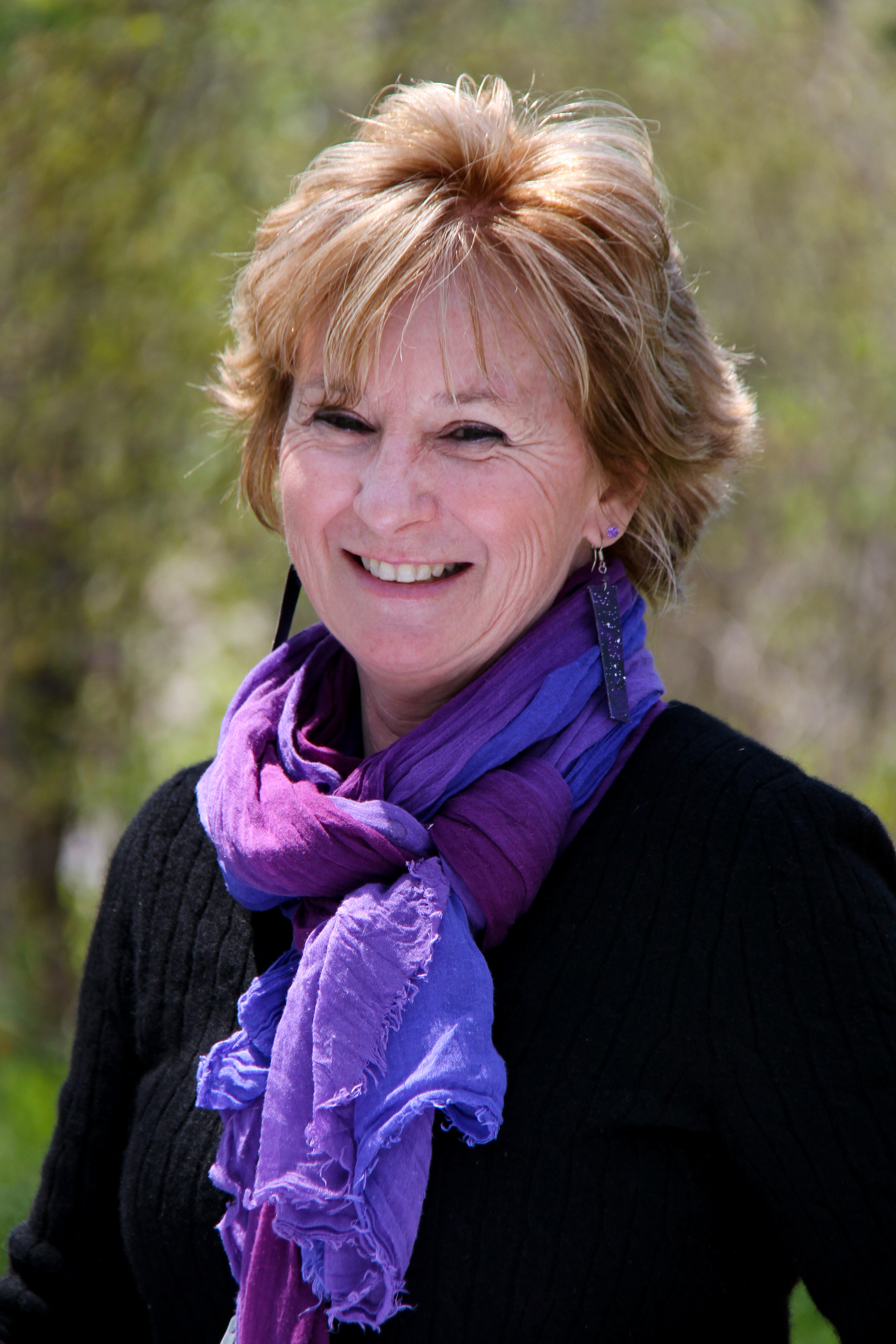 Peggy Rainville smiles at the camera on a sunny day in front of a background of blurry green trees and. She wears a black sweater and bright purple scarf and wears long, rectangular, dangling earrings.