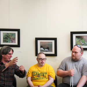 Three individuals are seated, facing the camera. They are engaged in conversation. The man at left gestures with his hand. They are seated in front of three framed photographs in black frames hanging on a white wall.