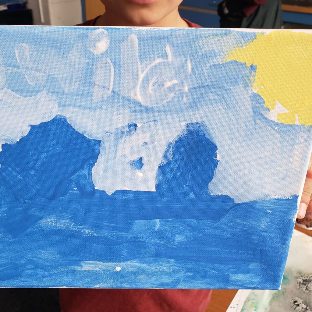 A student holds up a piece of artwork, covering their face and body. The painting is on a canvas, made up varying shades of blue