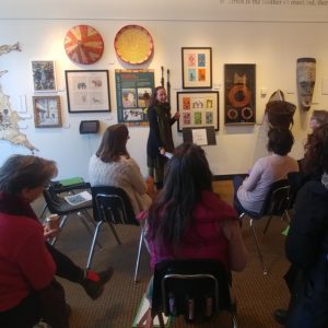 Alexandra Turner stands facing a group of six teaching artists, seated with their backs to the camera. She gestures to the wall behind her, filled with artwork from an exhibition at the Flynn Center.