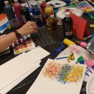 Viewed from above, a table is covered in art supplies including paint, scissors, paper, glue, brushes, and adaptive materials. A hand enters from left, dipping a brush into a palette of watercolors.