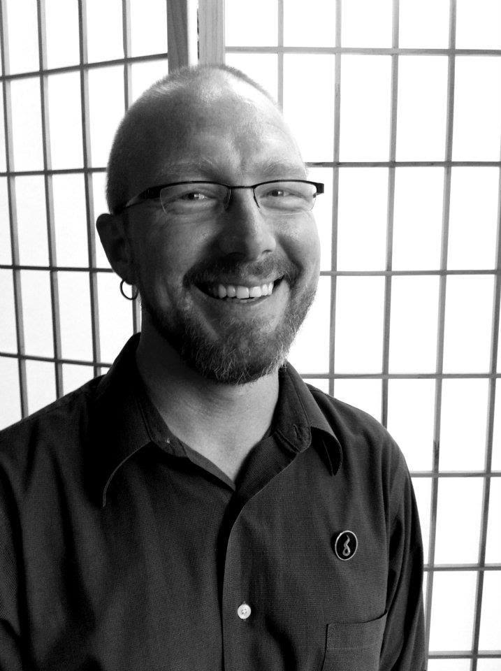 In a black and white photograph, Scott is seated in slight profile although his eyes and smile are directed to the camera. His hair and beard are closely trimmed. He wears dark metal framed glasses, small hoop earrings, and a dark button down shirt that is open at the collar.