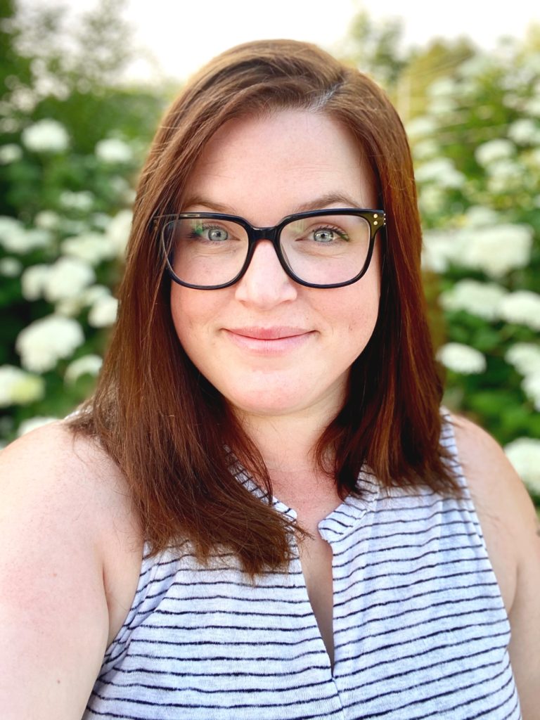 Katie Miller looks at the camera from behind black glasses. She has auburn hair and wears a white and black striped shirt. She stands in front of a bush of whit hydrangea blooms.