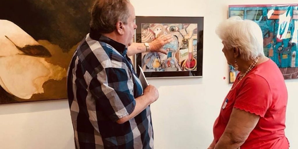 Two people stand in front of a piece of art. The person on left is pointing to a detail in the piece and speaking as they describe the detail.