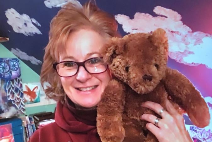 Peggy smiles warmly at the camera. She is in a room with slanted ceilings that are painted like the sky. She holds a brown teddy bear. Behind her. books and art are on shelves. Also, a painting of an owl peaks out from the left side of the photo.