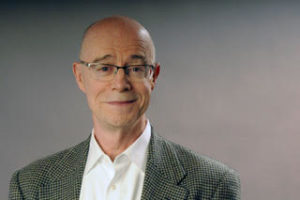 John Killacky stands in front of a gradient background that transitions from dark to light gray. He has pale skin, blue eyes, and is balding. He wears a pair of gray and clear glasses and a gray, checked blazer over a white button-down shirt. 