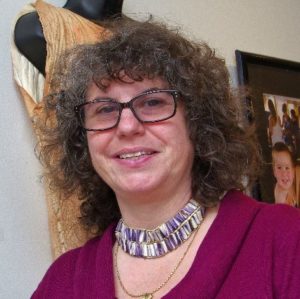 This is a headshot/picture of Vera, who has brown hair, brown eyes, and is wearing brown glasses. She is standing in front of a white wall with a framed woven textile hanging on the wall.