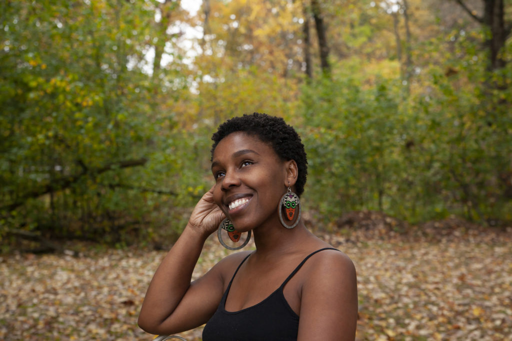 janet e. dandridge is sitting on an angle, looking towards the sky with a fall landscape surrounding her. She is wearing hand-made hoop earrings, and a black tank top. janet has dark-brown skin and eyes, and sports a short, dark brown tightly curled afro.