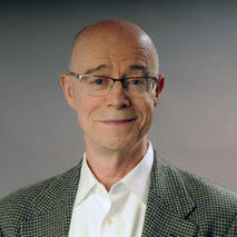 John Killacky stands in front of a gradient background that transitions from dark to light gray. He has pale skin, blue eyes, and is balding. He wears a pair of gray and clear glasses and a gray, checked blazer over a white button-down shirt. 
