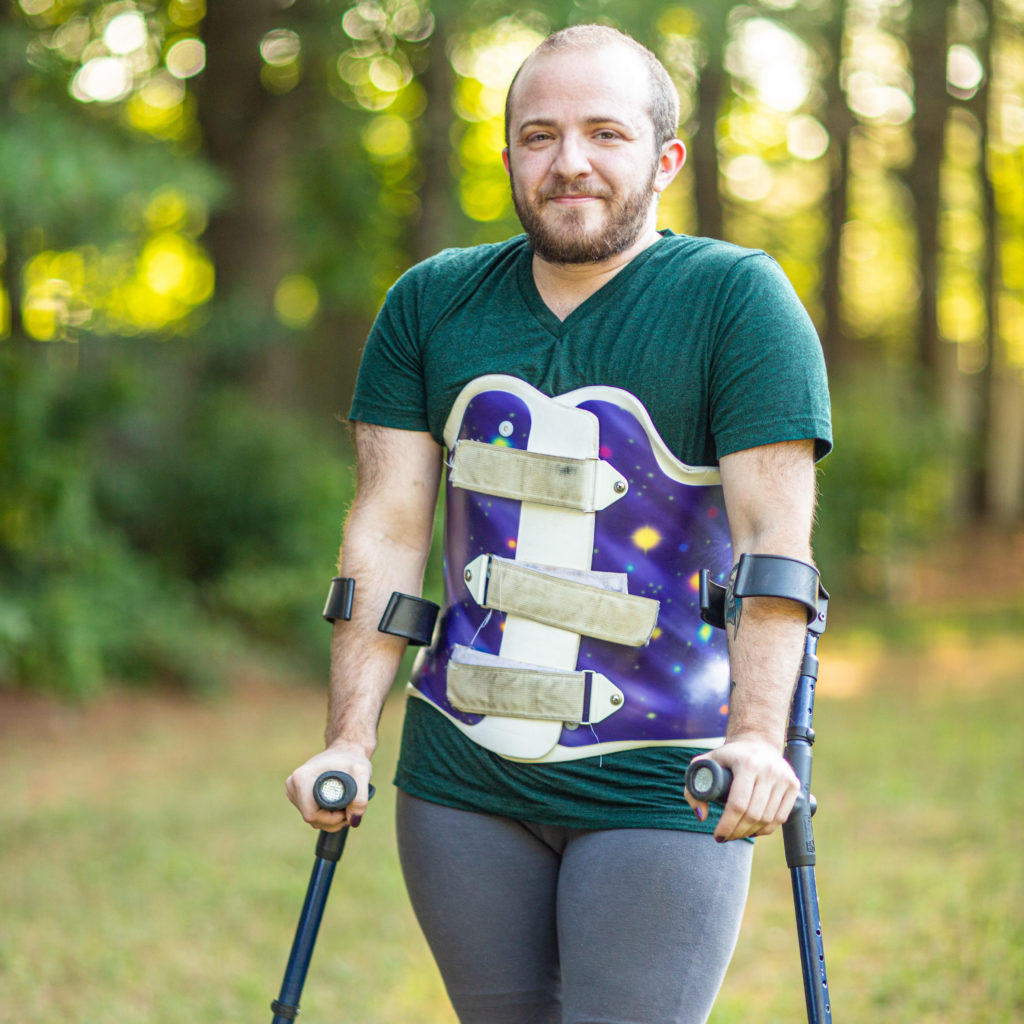Toby, a white person with a short dark beard and a shaved head, standing casually with forearm crutches, in leggings, t-shirt, and a purple galaxy print back brace. They are outdoors with soft-focus golden-green trees behind them.