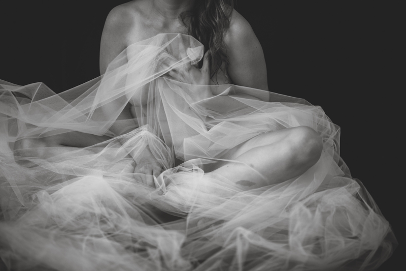 Fibrosis (Covered in Tulle) is a 20” by 30” horizontal, black and white, digital photograph. It features the seated, nude body of a woman wrapped in tulle against a black background. The fabric wraps and crinkles around the torso and legs, which are folded into a butterfly pose.