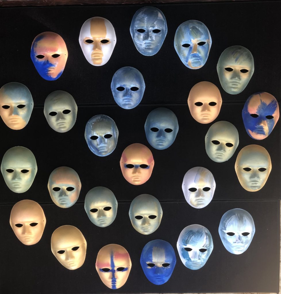 This piece is made up of painted papier-mache masks mounted on a large square of black foam core adhered to wood. The masks are all similar in shape, either child or adult size, with open almond-shaped eye holes, an angular, pointed nose, and a closed mouth in a neutral expression. There are 25 masks, arranged in five rows, or arcs. The masks are painted in combinations of orange, chrome, grey, gold, cobalt, light blue, and sage green. 