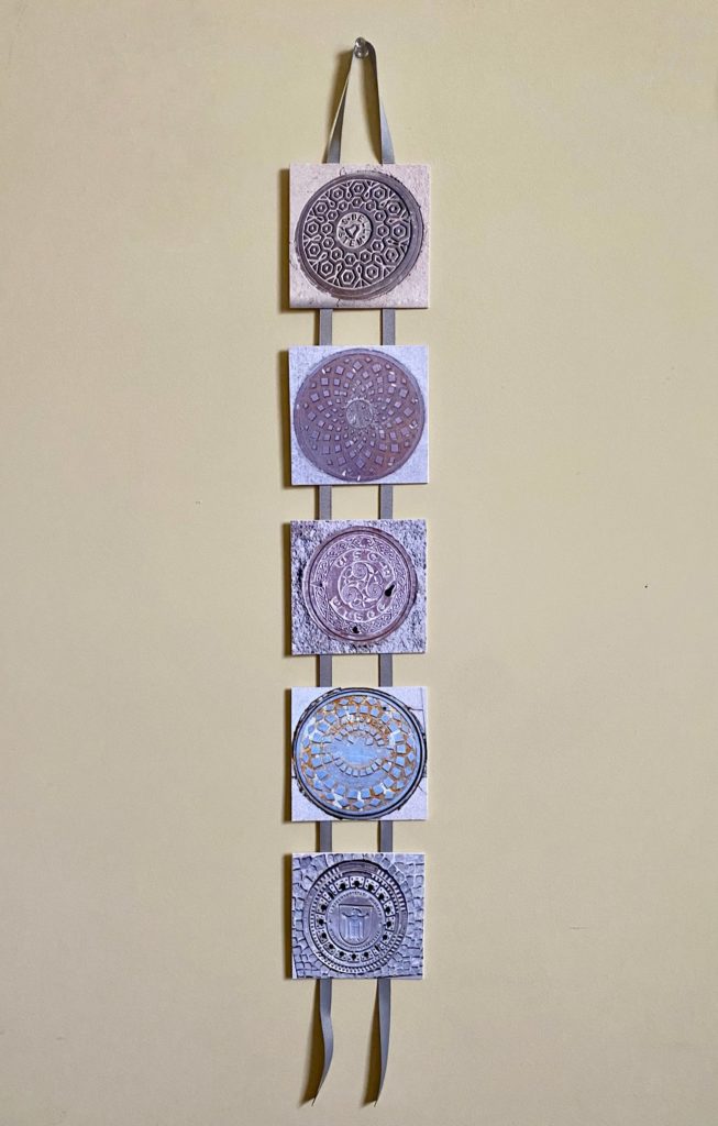 This is a collection of five square, color photographs, each 4 inches square, mounted in a ladder formation on gray ribbon. Each image is of a different manhole cover viewed from above.      The ribbon binding all of the images together is double the length of the piece and folded over to form a long u-shape at the top of the ladder, with two four-inch tails of ribbon coming out from the bottom. 