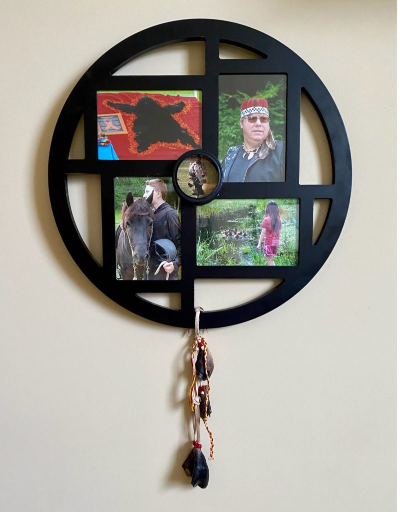 This photographic collage is 17 inches wide by 25 inches tall.  It features 4 rectangular black framed photos within a large black circular frame.  In the center of the 4 rectangular framed photos is a small photo in a circular frame. Open spaces within the circular frame are empty.  Hanging from the bottom of the circular frame is a white wide cord with knots, red beads, and four deer hooves attached. Also hanging from the frame is a braid of red, yellow, and white strings. 