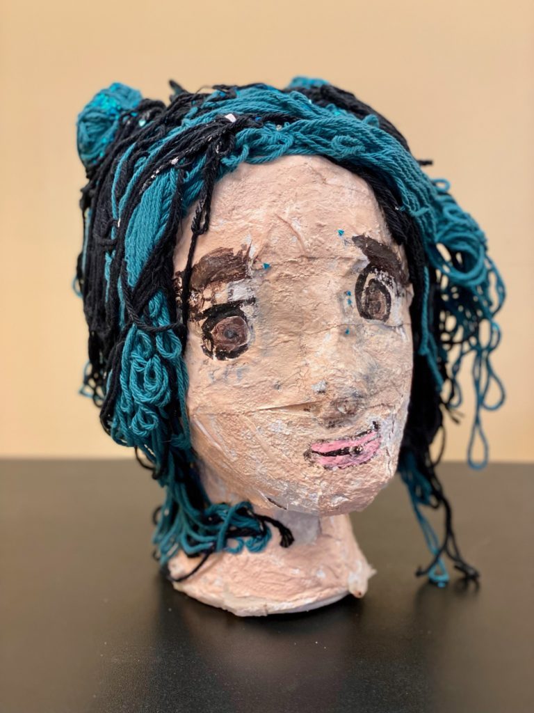 This sculptural work is of a person’s head and neck. It is constructed out of paper mache and foam, and features decorative elements of paint, yarn, and glitter. The skin is a pale beige, the eyes large and brown, and the hair is teal and black. 