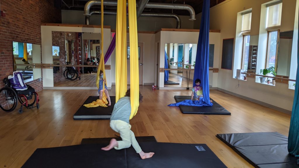 A photo of an aerial studio. There are three aerial silks, each with a child interacting with them. In the foreground, a child is mid-motion, with their feet dangling. In the background, two children look on. A wheelchair, mirrors, and mats are also in the frame.