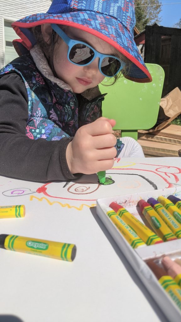 A child draws with oil pastels on white paper. The child is outside and wears a dark long-sleeved shirt, sunglasses, and a hat. There are oil pastels in a container and scattered around the child's creation.