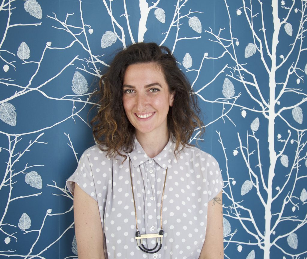 Abbey stands in front of a wall with blue and white wallpaper with tree branches and leaves in the pattern. Abbey has curly, shoulder length, brown hair, pale skin, and a bright smile. She's wearing a neutral polka dot top with a dangling metallic necklace with a large pendant.