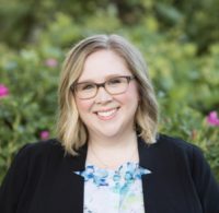 Kassandra is photographed in front of green foliage, with little peaks of yellow and pink flowers behind her. Kassandra wears glasses, has pale skin, and a blonde bob swept to the side. She smiles brightly at the camera. She wears a black blazer over a printed blue shirt.