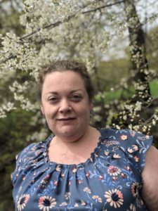 Masha stands in front of a flowering tree. She has light curly hair pulled back, pale, skin, and smiles softly at the camera. Masha wears a blue floral short-sleeved shirt.