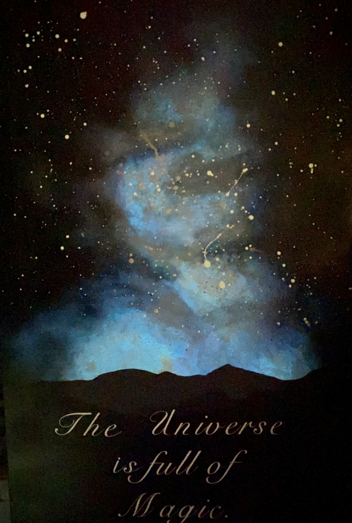 This is a 36” tall and 24” wide painting depicting a landscape scene of a far off mountain range at twilight. The top two-thirds of the painting make up the sky. The sky is vignetted in black, and fades into shakes of bright blue, lavender, and white that form a nebula-like center. Stars dot the sky, make up white paint dripped and flicked onto the canvas. This piece glows in the dark, and under the right conditions the blue portions of the painting glow. At the bottom a mountain range cascades into the background in layers of black, charcoal, and light gray. There is white scripted text over the mountains that reads “The Universe is full of Magic.”