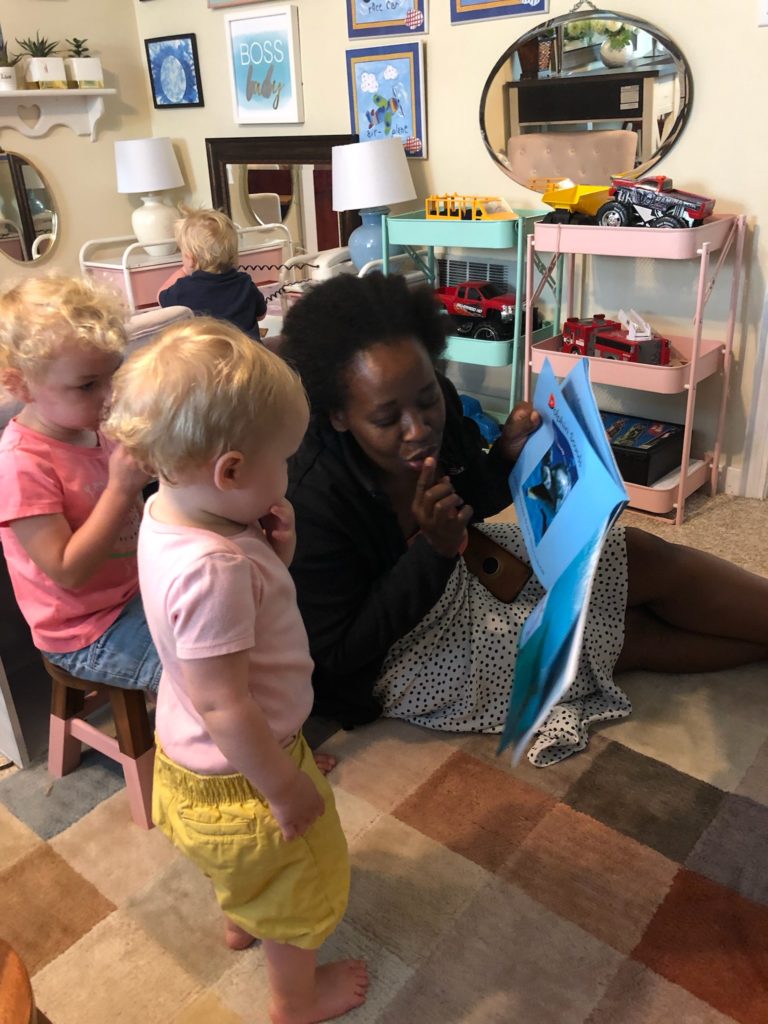 A childcare provider kneels down while reading a book to small children.