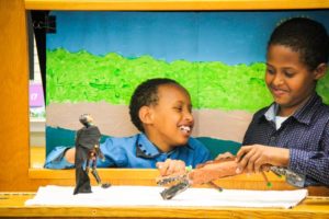 Two students put on a show behind a puppet theater. Students have a hand-painted landscape behind them and handmade puppets they use to put on a show. Both students smile.
