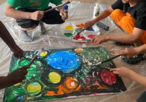 Students works together on a painting. They are seated or kneeling on the floor and painting with yellow, green, orange, red, and blue. The canvas has a dark green background and large, colorful orbs.