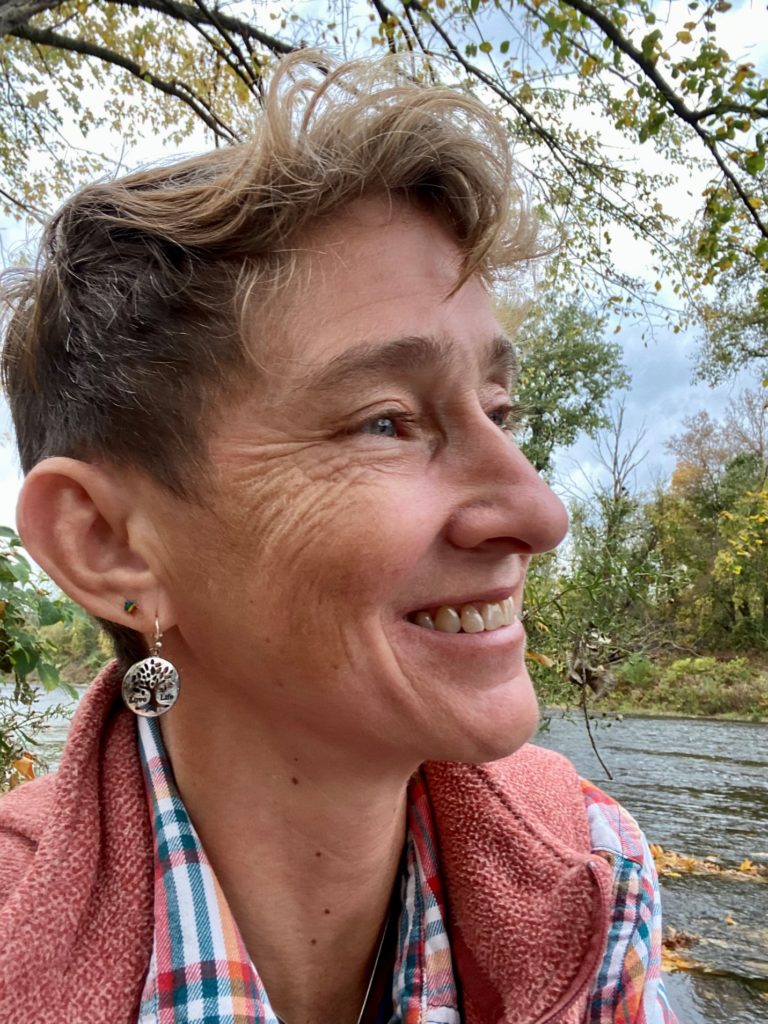 A headshot of Heidi Swevens, she/they, with blue eyes, pale skin, and short brown hair. They smile, looking to the side, wearing a plaid shirt and coral fleece vest with a river and trees in the background.