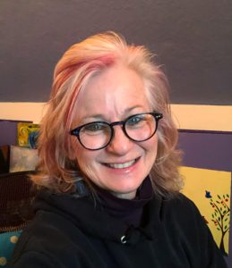 Peggy smiles at the camera. She has pale skin, blonde hair with a red streak, and wears a black turtleneck and black glasses. She's sitting in a home office with paintings behind her and tree murals on the walls.