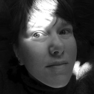 A black and white portrait of Megan Bent. She has pale skin and dark bangs sweeping over her forehead. Most of her face is in a light shadow and the sun illuminates one eye.