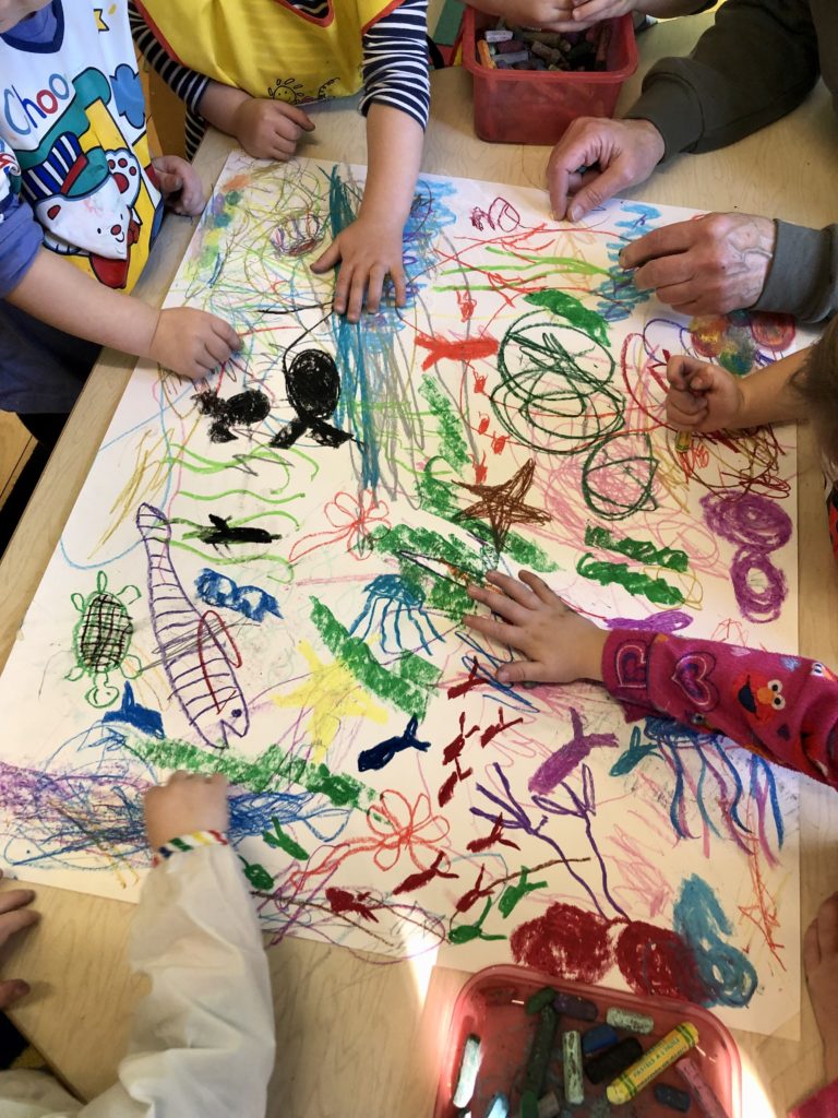 Children create a collaborative picture, based on the book Swimmy, featuring all sorts of aquatic life. Viewed from above, children's hands use a variety of implements to add to the picture.