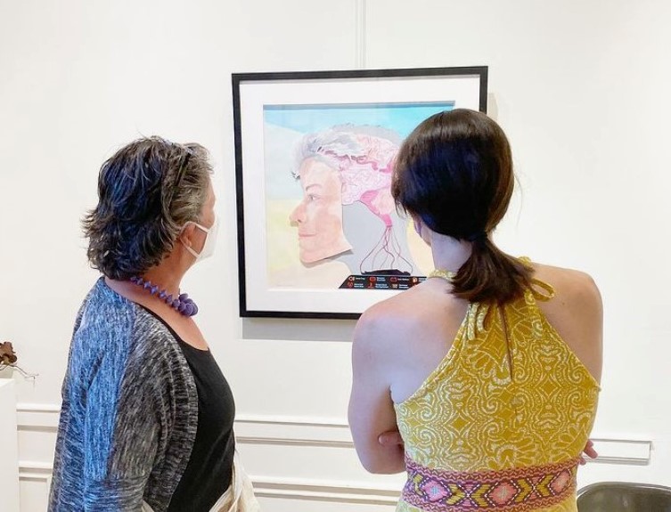 Two people stand in front of a piece of art by Lissa Nilsson. The piece shows Lissa's head in profile, pale skin, grey hair. The brain is shown and is mapped with side effects of chronic pain and illness. It's displayed on a gallery wall.