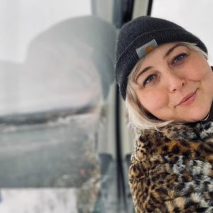 Kat sits inside a gondola over water. She leans her head against the glass, her reflection mirroring her pose. Kat wears a grey Carhartt hat and a fuzzy leopard print jacket. She smiles, round cheeks lifting. Kat has pale skin, light eyes, ans platinum blonde hair