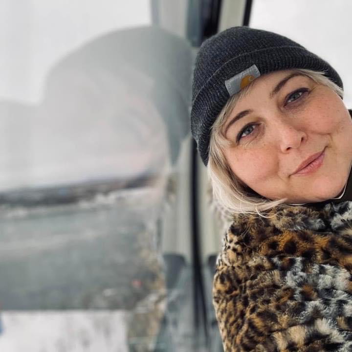 Kat sits inside a gondola over water. She leans her head against the glass, her reflection mirroring her pose. Kat wears a grey Carhartt hat and a fuzzy leopard print jacket. She smiles, round cheeks lifting. Kat has pale skin, light eyes, and platinum blonde hair
