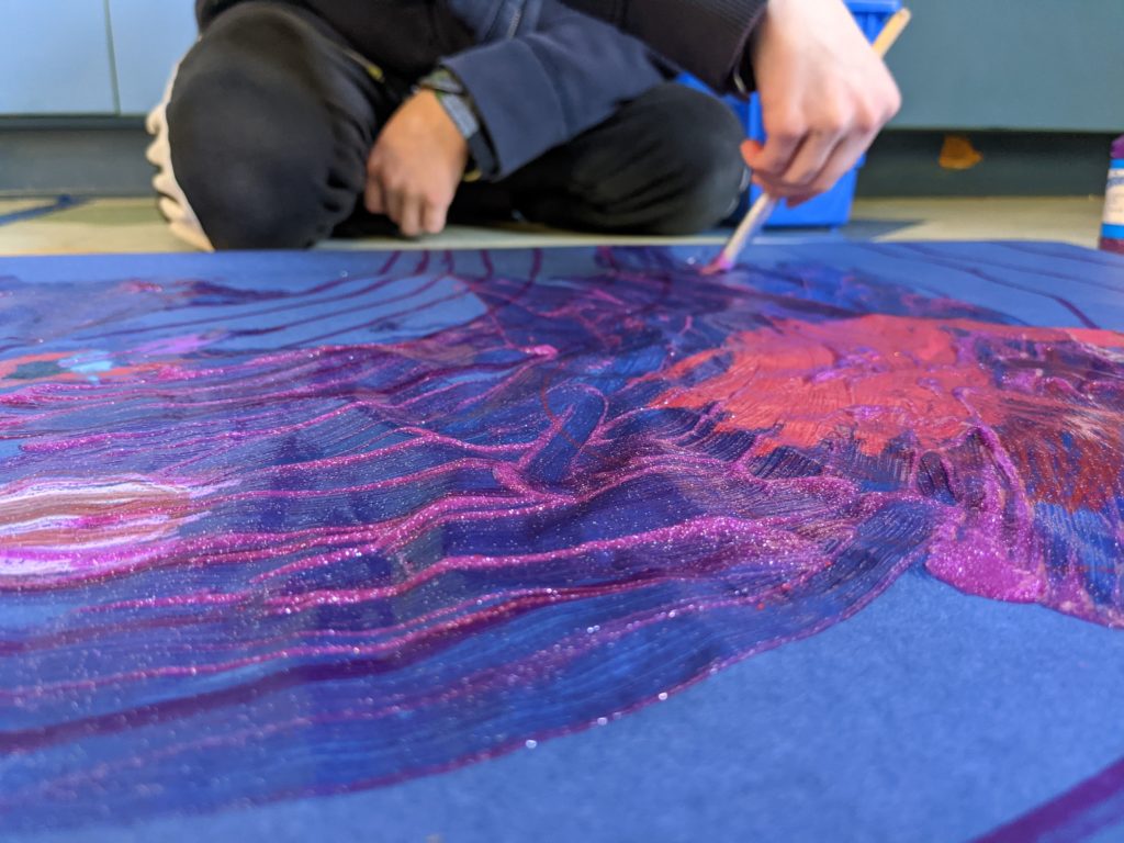 A student sits on the floor cross-legged and paints on blue paper with sparkling purple, blue, pink, and white paint.