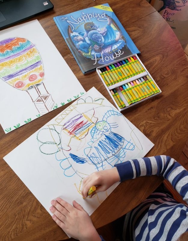 A child sits at a table drawing with oil pastels. On the table is a picture drawn by a parent, a box of oil pastels, and the book, The Napping House.