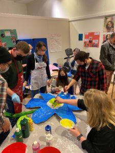 Students and teachers work collaboratively to paint giant flowers to add to their pollinator installation project. The flower is bright blue with a yellow center.