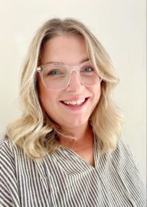 Katie Miller looks at the camera from behind clear glasses. She has pale skin, shoulder-length blond hair, and is wearing a grey and white striped shirt. She is smiling and standing in front of a beige wall.