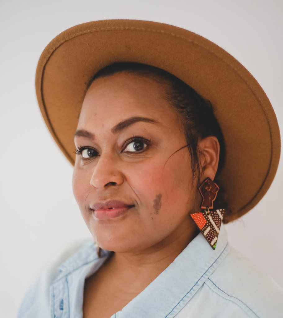 A headshot of Jen White-Johnson. Jen is shown from the shoulders up, with her head turned towards the camera. Her hair is pulled back and she wears a camel wool hat. Her earrings are wooden, a brown fist attached to a triangle with white, coral, and green segments and tan dots. She wears a pale denim collared shirt. Jen has brown skin, deep brown eyes, and rosy cheeks.