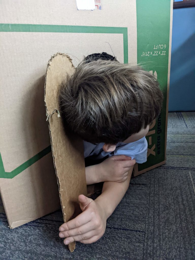 A student peaks their head out from an opening in a cardboard box. The opening is a door to their own classroom tiny house.