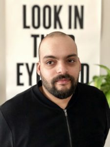 A headshot of Jeff Kasper. Jeff is shown from the chest up, looking straight at the camera. He is wearing a black zip-up cardigan, and behind him is an out of focus artwork composed of black text on a white ground. The only words visible from the text is the phrase “look in”. Jeff has brown skin, brown eyes, and a tightly-shaven haircut, mustache, and beard.