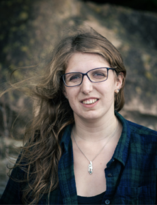 A vertical portrait of Aurora a woman with pale skin, long blond hair, and black-rimmed glasses. She is wearing a blue and green flannel shirt and is outdoors with trees behind her.