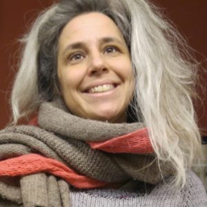 A pale woman with a welcoming smile, lively brown eyes, and a rambunctious river of long gray hair beams at the camera with her head tilted. She is enveloped in layers of hand knits. 