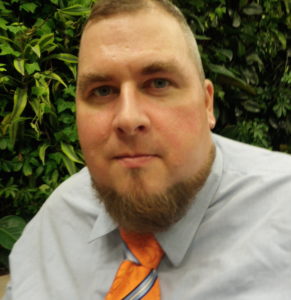 Portrait of Jason Angel. Jason is a white male with blond hair, blue eyes, and a beard. He is wearing a light blue dress shirt and an orange tie. There is foliage behind him.