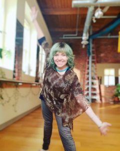 Portrait of Jennifer in a large loft space with brick walls and windows. Jen has pale skin, green hair, and a brown long-sleeved shirt and grey pants. She is leaning an arm on one knee and smiling.