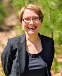 A portrait of Sarah. She has pale skin, short red hair and is smiling at the camera. She is wearing glasses a black tank top and a blazer. There is green foliage behind her. 