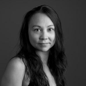 A black and white headshot of Maya. Maya is Japanese-American with long brown hair, light skin, and is wearing a black tank top. She is smiling towards the camera and is in front of a grey background.